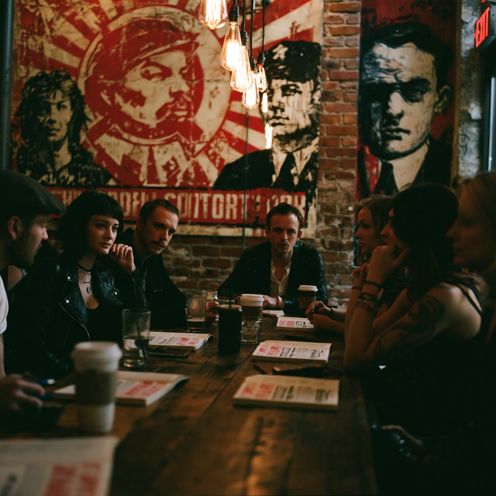 A book club of five around a communal table, identical unopened paperbacks beside their drinks